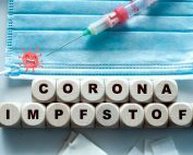 auf weißem Tisch liegen Würfel mitz dem Schriftzug Corona Impfstoffe, eine Maske und eine Spritze mit weindendem Coronavirus, Credit: Canva